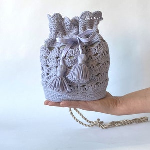 Puede incluir: Una bolsa de cordón de crochet lavanda con una cinta blanca y dos borlas. La bolsa tiene una correa de cuerda beige.