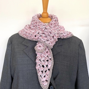 Puede incluir: Bufanda de ganchillo hecha a mano en tonos rosa, blanco y gris, expuesta sobre un maniquí de madera. La bufanda tiene un diseño de tejido abierto y texturizado con un borde gris liso. La bufanda está drapeada sobre una blazer gris.