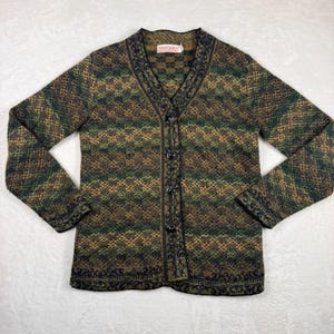 May include: A vintage cardigan sweater in shades of green, brown, and tan, featuring a patterned design. The sweater has a V-neck, long sleeves, and a button-up front. The label reads "INDO CARLO".