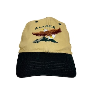 Puede incluir: Una gorra de béisbol de color tostado con visera negra que presenta un gráfico bordado de un águila calva volando sobre una cordillera de color verde oscuro. La palabra ALASKA está bordada en letras verde oscuro sobre el águila.
