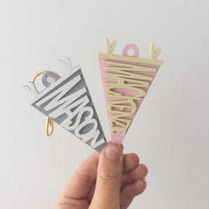 May include: Two pennant-shaped keychains, one gray and one pink, each with a name cut out in a contrasting color. The gray keychain says "MASON" and has a gold-colored keyring. The pink keychain says "MCKENZE".