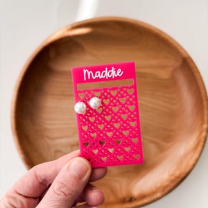 May include: A bright pink earring display card with a heart-shaped cut-out design and the name "Maddie" in white script. Two white pearl earrings are displayed on the card. The card is held in front of a wooden bowl.