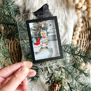 May include: A clear acrylic ornament with a black frame holding a photo of a baby in front of a Christmas tree. The ornament has the text "The Bell Family" written on it.