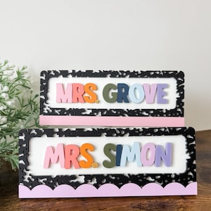 May include: Two rectangular desk signs with the names "MRS. GROVE" and "MRS. SIMON" in colorful 3D letters. Each sign has a black and white patterned frame, a white interior, and a pink scalloped base.