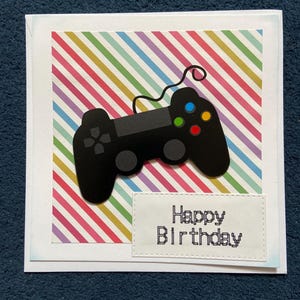 May include: A square greeting card featuring a black video game controller graphic centred over a background of diagonal multi-coloured stripes. A white rectangular label with stitched edges displays the text Happy Birthday in black pixelated font.