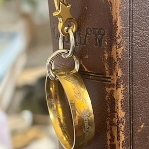 May include: A gold-colored ring with engraved text, attached to a gold keychain with a star-shaped charm. The ring is suspended in front of a worn, brown leather-bound book.