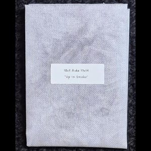 May include: A folded piece of light gray 18ct Aida fabric, measuring 19x14 inches, with the text "Up In Smoke" on a white label. The fabric has a textured appearance, and the background is dark.