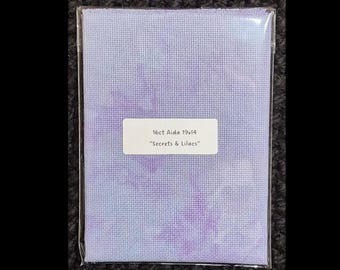 Pode incluir: Tecido Aida para ponto cruz, embalado em plástico transparente. O tecido é uma mistura de tons de lavanda e roxo. Uma etiqueta branca diz "16ct Aida 19x14" e "Secrets & Lilacs."