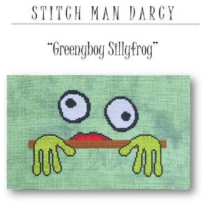 May include: A cross-stitch design featuring a cartoon frog with large eyes, a red mouth, and green hands. The frog is holding a brown object. The text "Greenyboy Sillyfrog" is above the design, and "Stitch Man Darcy" is at the top.