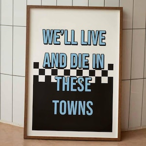 Może przedstawiać: Oprawiony wydruk artystyczny z napisem "WE'LL LIVE AND DIE IN THESE TOWNS" w jasnoniebieskim na białym tle. Projekt zawiera czarno-biały wzór w szachownicę i czarny prostokąt. Rama jest brązowa.