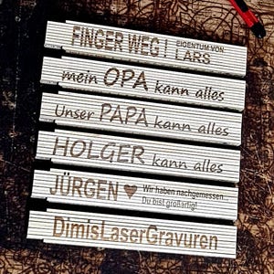 Könnte beinhalten: Eine Sammlung von Holzgliedermaßstäben mit Gravuren. Die Lineale zeigen Sätze wie "FINGER WEG!" und Namen wie "LARS" und "HOLGER". Die Lineale sind wahrscheinlich personalisierte Geschenke, mit dunkelbraun geätztem Text.