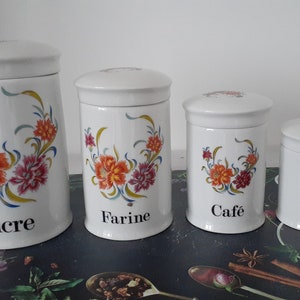 Peut inclure: Ensemble de quatre pots de cuisine en céramique blanche avec couvercles, décorés de motifs floraux orange, rose et bleu. Les pots sont étiquetés "sucre", "farine", "café" et "thé".