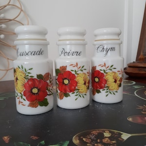 May include: Three white ceramic spice jars with floral designs. The jars have black lettering that reads "Cuscade", "Poivre", and "Chym".