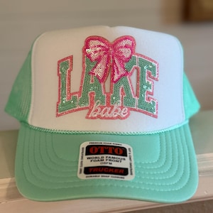 May include: A mint green and white trucker hat with the words "LAKE babe" in sequined letters and a sequined bow. The hat has a curved brim and a mesh back. The hat has a tag that says "OTTO WORLD FAMOUS FOAM FRONT TRUCKER".