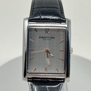 May include: A rectangular Kenneth Cole New York watch with a silver face and black leather strap. The watch face has rose gold hands and markers. The strap is black and has a textured, crocodile-like pattern.