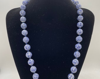 May include: A necklace featuring white and blue ceramic beads with a repeating geometric pattern. The beads are strung together on a blue cord, creating a decorative accessory. The necklace is displayed on a black mannequin.