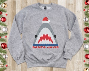 May include: A gray sweatshirt with a graphic of a shark wearing a Santa hat. The text "SANTA JAWS" is printed below the shark.