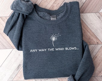 May include: A dark gray sweatshirt with a white dandelion graphic and the text "ANY WAY THE WIND BLOWS..." printed on the front.