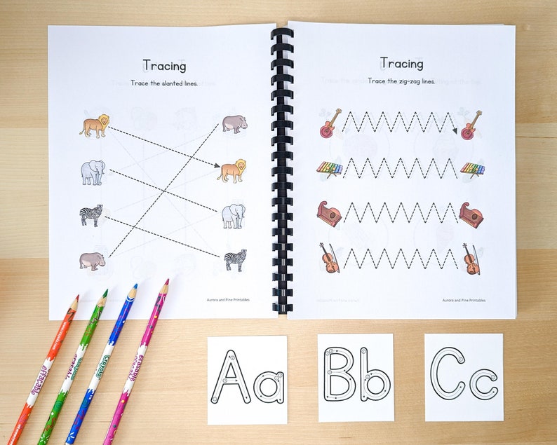 May include: An open workbook with tracing activities for kids. The left page features a tracing activity with animals, while the right page has a tracing activity with musical instruments. The workbook is open to show the pages and a set of colored pencils are lying on a wooden surface. Three alphabet flashcards are also visible on the surface, showing the letters A, B, and C.