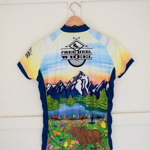 Pu&ograve; includere: Una maglia da ciclismo blu, gialla e bianca con una scena di montagna e un alce. La maglia ha il testo "Free Heel and Wheel" sul retro.