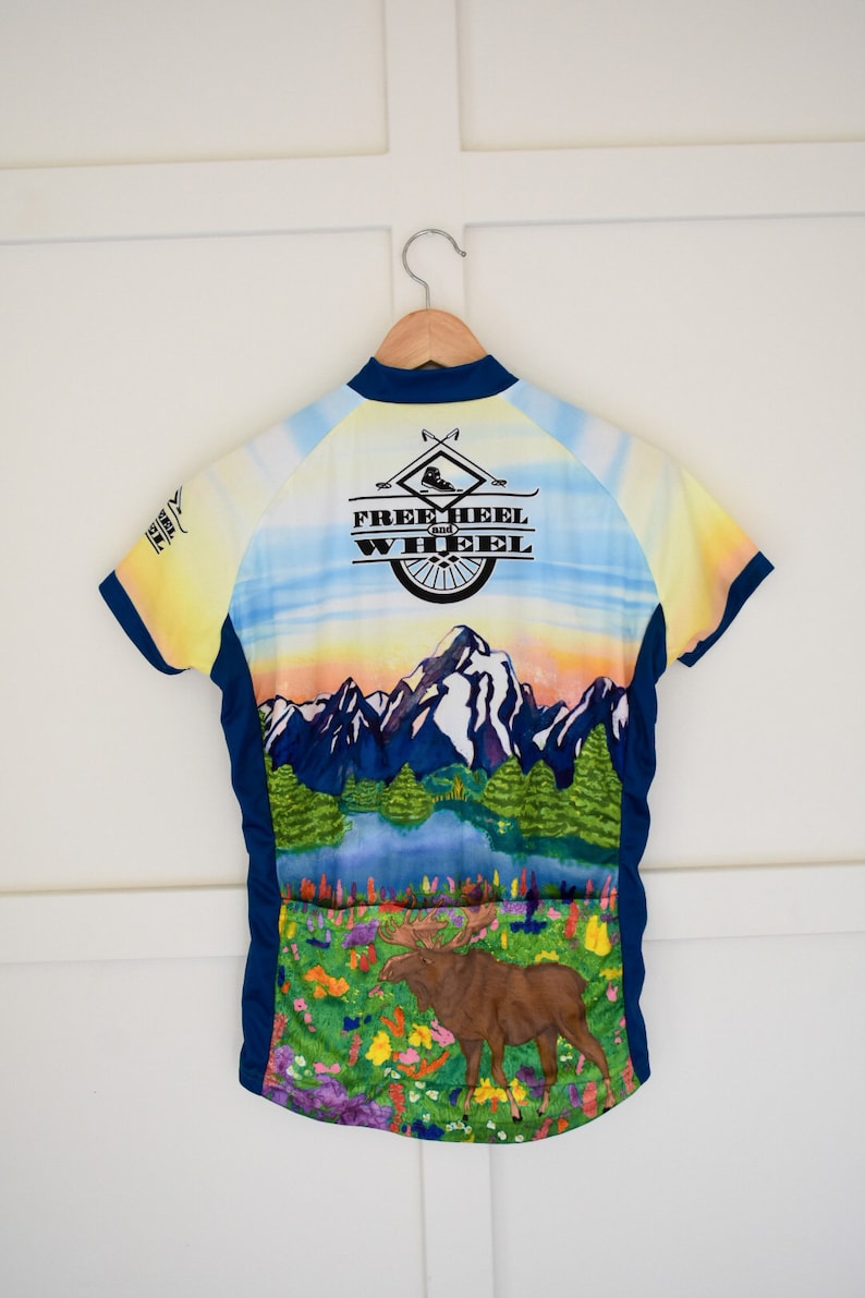 Pu&ograve; includere: Una maglia da ciclismo blu, gialla e bianca con una scena di montagna e un alce. La maglia ha il testo "Free Heel and Wheel" sul retro.