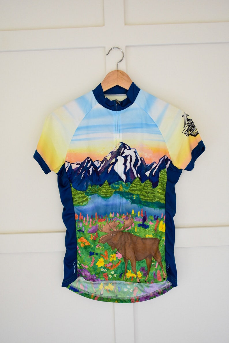 Pu&ograve; includere: Una maglia da ciclismo blu, gialla e verde con una scena di montagna e un alce. La maglia ha una cerniera a tutta lunghezza e maniche corte.