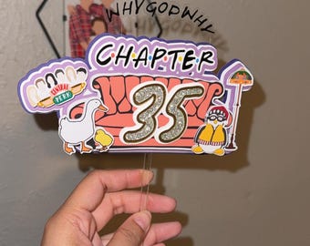 May include: A colorful cake topper with the text "CHAPTER 35" and a glittery number 35. The design includes cartoon elements like a couch, a duck, and a penguin, with the Central Perk logo. A photo is in the background.