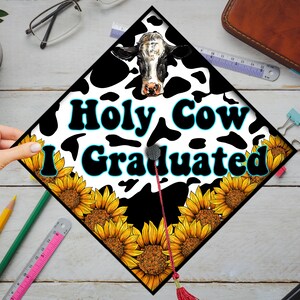 Może przedstawiać: Czarno-biały czepek absolwenta z nadrukiem krowiej sk&oacute;ry z słonecznikami i tekstem "Holy Cow I Graduated" w kolorze białym i niebieskim.