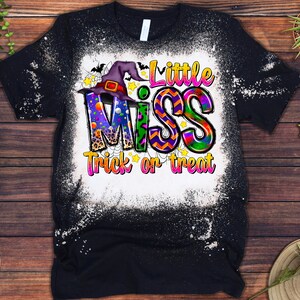 May include: Black t-shirt with a distressed white design. The shirt features the text "Little Miss Trick or Treat" in colorful, playful lettering, with a witch hat and stars. A fun Halloween-themed graphic tee.