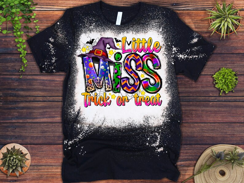 May include: Black t-shirt with a distressed white design. The shirt features the text "Little Miss Trick or Treat" in colorful, playful lettering, with a witch hat and stars. A fun Halloween-themed graphic tee.