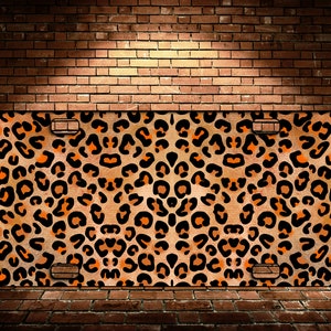 May include: A brown and orange leopard print license plate cover. The cover has black spots and a slightly textured surface.