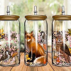 Puede incluir: Tres frascos de vidrio con tapas y pajitas, cada uno con un animal del bosque y un diseño floral diferente. Los frascos presentan un zorro, un búho y un pájaro, rodeados de flores rosas, hojas marrones y un estampado de leopardo.