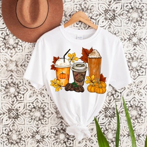 May include: White t-shirt with a graphic design of three fall-themed coffee drinks, including a pumpkin spice latte, a regular coffee, and a caramel macchiato. The design also includes fall leaves, pumpkins, and coffee beans.