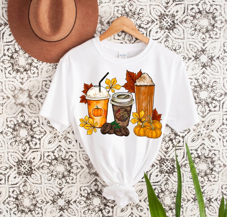 May include: White t-shirt with a graphic design of three fall-themed coffee drinks, including a pumpkin spice latte, a regular coffee, and a caramel macchiato. The design also includes fall leaves, pumpkins, and coffee beans.