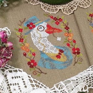 May include: Cross-stitch embroidery of a goose wearing a blue hat and a red bow tie, surrounded by a wreath of red, orange, and yellow flowers. The background is a beige linen fabric.