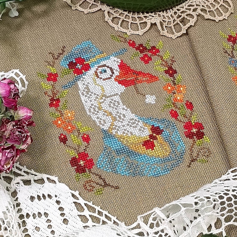 May include: Cross-stitch embroidery of a goose wearing a blue hat and a red bow tie, surrounded by a wreath of red, orange, and yellow flowers. The background is a beige linen fabric.