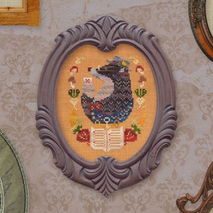 May include: A framed cross-stitch embroidery featuring a brown and black bear wearing a blue and white hat and holding a book. The bear is surrounded by flowers, mushrooms, and other nature-inspired elements. The embroidery is set in a dark brown oval frame.