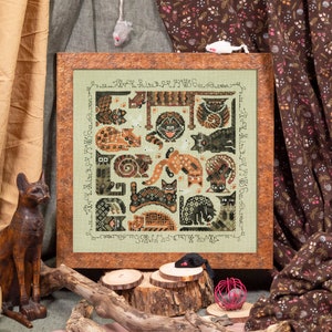May include: A framed cross-stitch design featuring a variety of cats in different poses and patterns. The cats are stitched in shades of brown, black, and white on a green background. The frame is brown wood with a distressed finish.