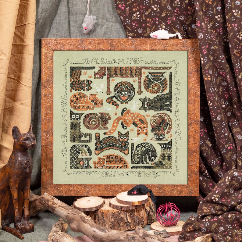 May include: A framed cross-stitch design featuring a variety of cats in different poses and patterns. The cats are stitched in shades of brown, black, and white on a green background. The frame is brown wood with a distressed finish.