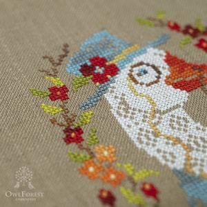 May include: Close-up of a white goose cross-stitch embroidery with a blue hat and red flowers. The embroidery is on a beige linen fabric.