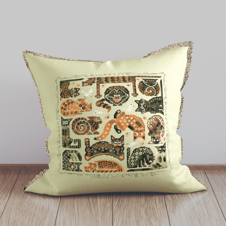 May include: A beige pillow with a brown and orange embroidered design of cats. The design features a variety of cats in different poses, including sleeping, playing, and sitting. The pillow is decorated with a brown and white trim.