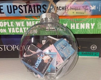 May include: A clear glass Christmas ornament filled with various book covers. The covers include titles like "Losers" by Harley Laroux and "Pucking Around".
