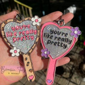 May include: Two heart-shaped keychains with the text "You're like really pretty." One keychain has a leopard print handle and white flowers. The other has a pink handle and purple flowers. Both have silver glitter.