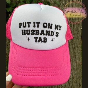 May include: Pink and white trucker hat with black text that reads "Put it on my husband's tab" with two small glitter stars on either side of the word "tab".