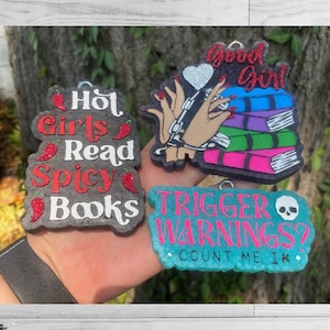 May include: Three resin book-themed charms with text. One reads "Hot Girls Read Spicy Books" with chili pepper accents. Another says "Good Girl" with a heart and books. The third says "Trigger Warnings? Count Me In" with a skull.