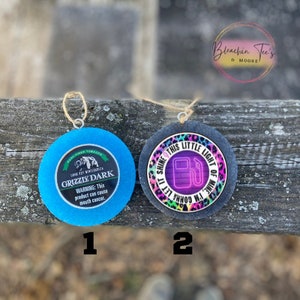 May include: Two round ornaments with different designs. The first ornament is blue with a white circle in the center that says "Grizzly Dark" with a bear image. The second ornament is black with a pink, purple, and green leopard print design and a neon pink gas pump with the text "Shine this little light of mine, I'm gonna let it shine".