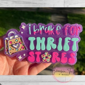 May include: A purple, glittery resin keychain with the phrase "I brake for thrift stores" in pink and teal lettering. The keychain features a shopping bag design with a heart and leopard print accents.