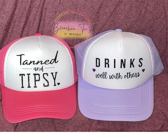May include: Two trucker hats with white mesh backs and different colored brims. The pink hat says "Tanned and Tipsy" in black text. The purple hat says "Drinks well with others" in black text with two small pink hearts.