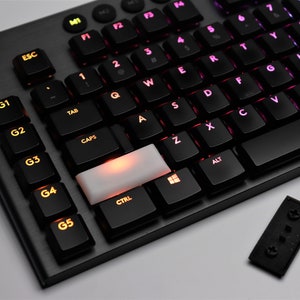 Könnte beinhalten: Eine schwarze Gaming-Tastatur mit einer weißen Tastenkappe und einem kleinen schwarzen USB-Empfänger. Die Tastatur hat ein G-Logo und G1-G5-Tasten auf der linken Seite. Die Tasten sind mit verschiedenen Farben beleuchtet.