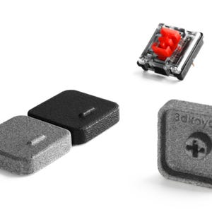 Puede incluir: Un interruptor de teclado mecánico con un vástago rojo y una carcasa transparente, acompañado de dos teclas cuadradas en negro y gris, y una tecla gris que muestra la parte inferior con el texto 3dkeycap y un soporte en cruz.